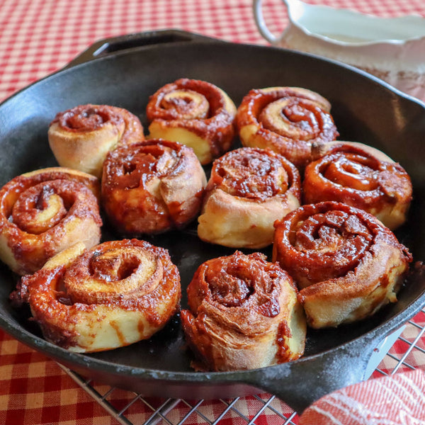 Baked cinnamon rolls in a cast iron skillet on a red checkered tablecloth.