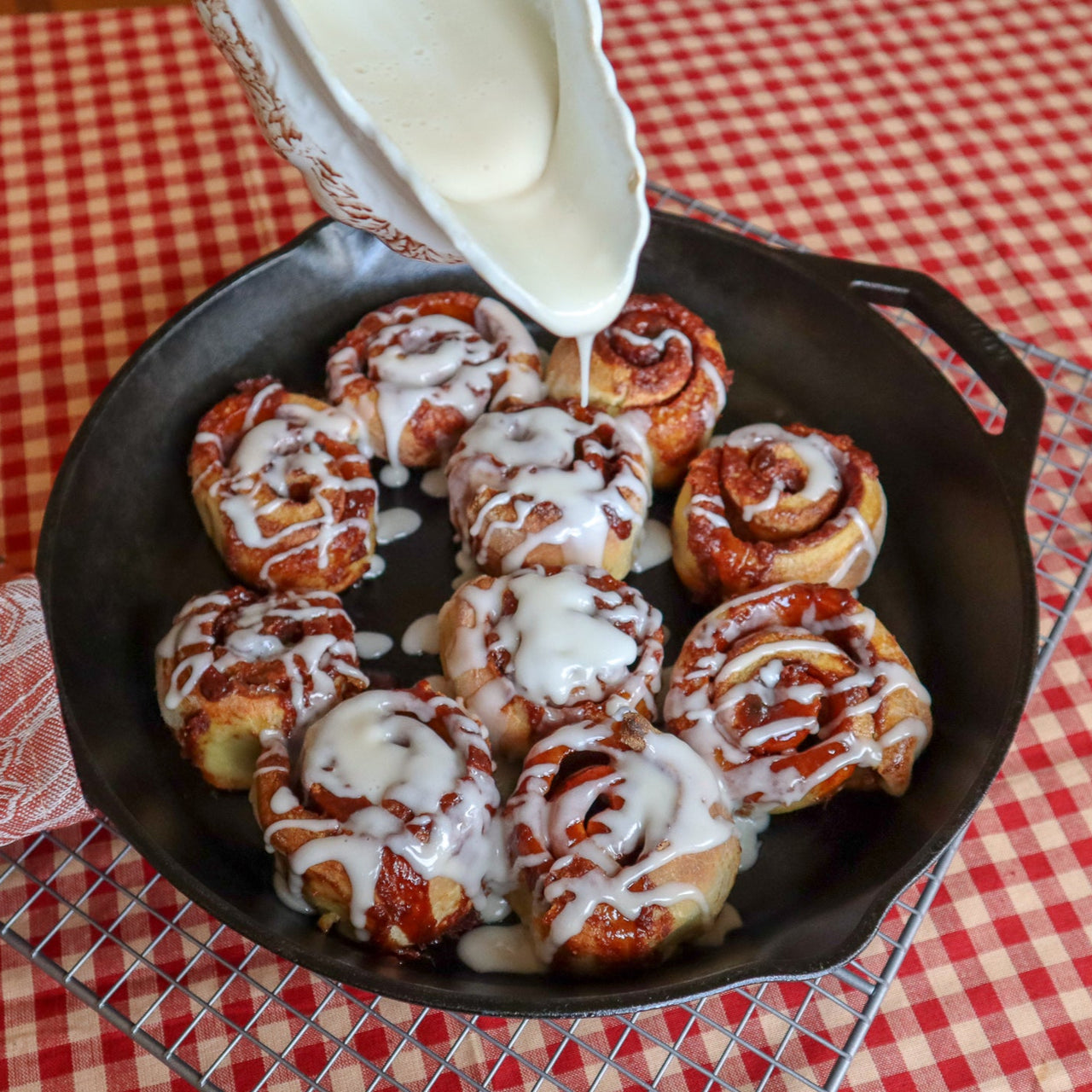 Cinnamon rolls with icing being poured over them in a black skillet on a red and white checkered tablecloth.